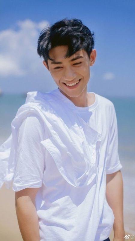 Người mặc áo trắng cười tươi đứng trên bãi biển, trời xanh và nắng đẹp.