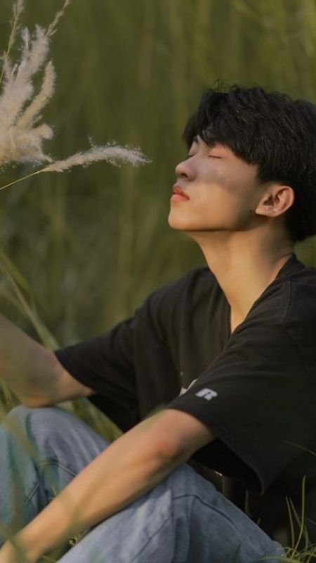 Người ngồi trong cỏ xanh, nhắm mắt thư giãn, ánh sáng chiếu nhẹ lên khuôn mặt, tay chạm cành lau.