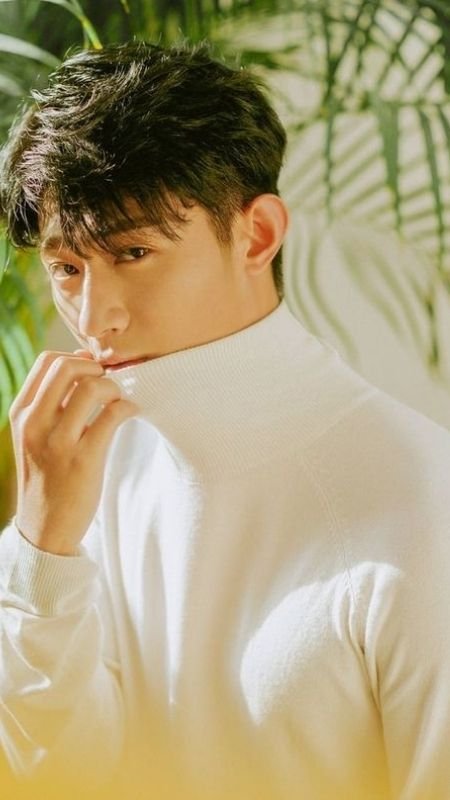 Người mặc áo len cổ lọ màu trắng, đứng gần cây xanh, ánh sáng chiếu sáng khuôn mặt và bối cảnh.