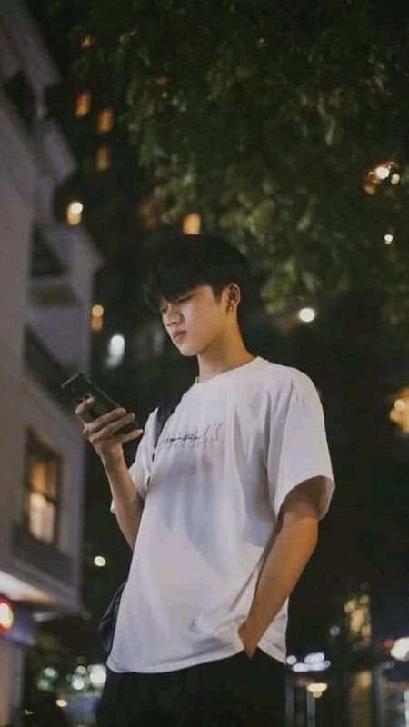 Người trẻ mặc áo phông trắng đứng ngoài trời vào ban đêm, tay cầm điện thoại, xung quanh là cây và ánh đèn.