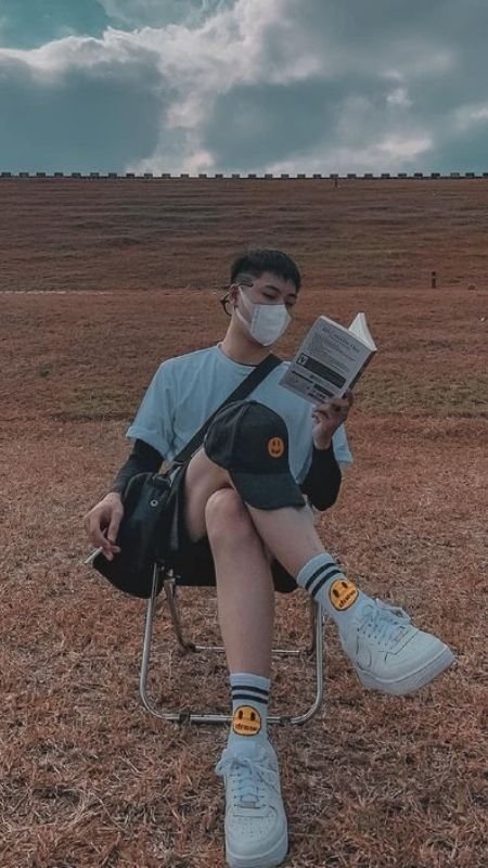 Người ngồi trên ghế đọc sách ngoài trời, đeo khẩu trang, mặc áo phông trắng, xung quanh là bãi cỏ rộng lớn.