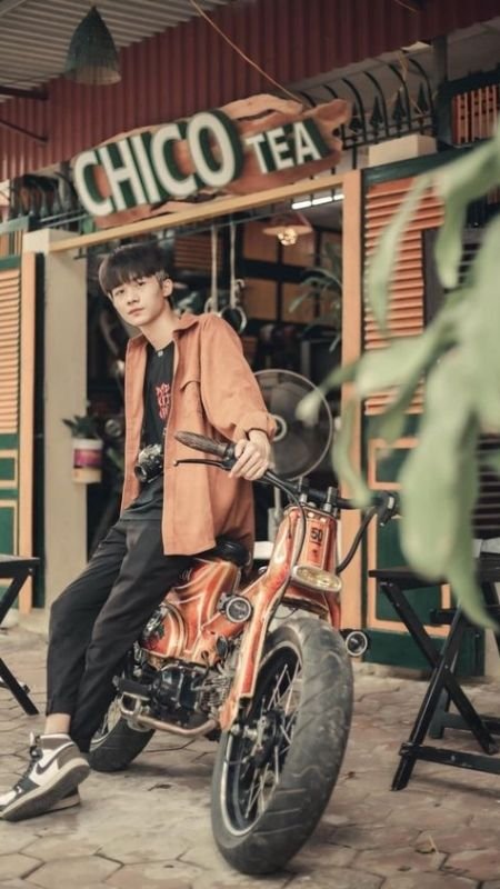 Người ngồi trên xe máy cổ điển trước cửa tiệm trà, xung quanh có bàn ghế và cây xanh.