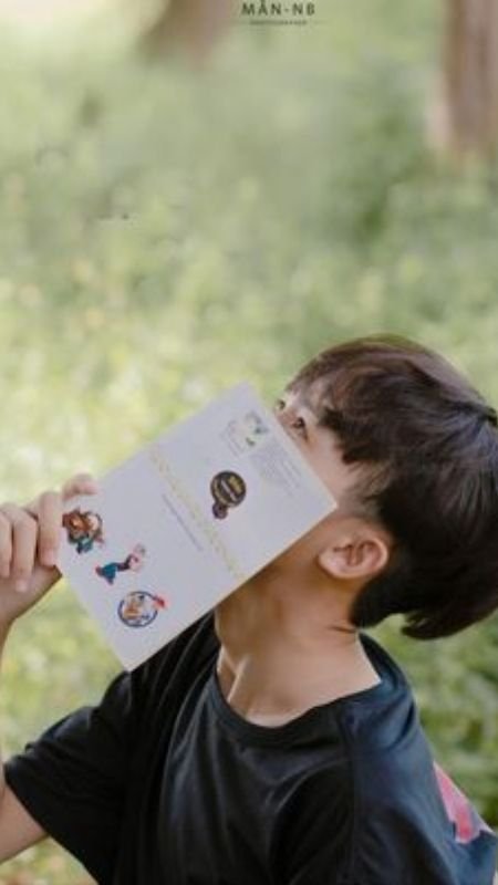 Người mặc áo đen cầm quyển sách che mặt, đứng trước nền cây cối xanh tươi.