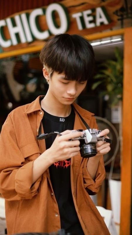 Người đứng cầm máy ảnh, mặc áo khoác màu nâu, nền có bảng hiệu quán trà.