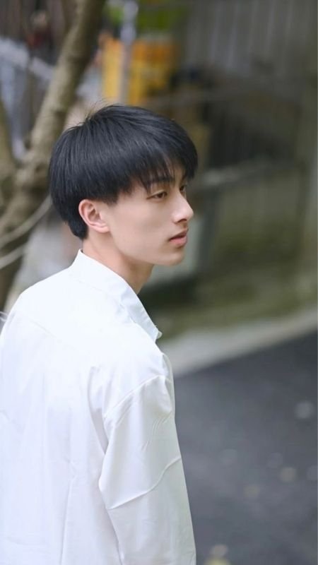 Người mặc áo sơ mi trắng đứng ngoài trời, quay đầu nhìn sang một bên, nền có cây và đường phố mờ nhạt.