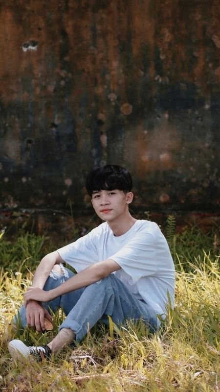 Người ngồi trên cỏ xanh, mặc áo trắng và quần jeans, dựa lưng vào bức tường cũ màu nâu.