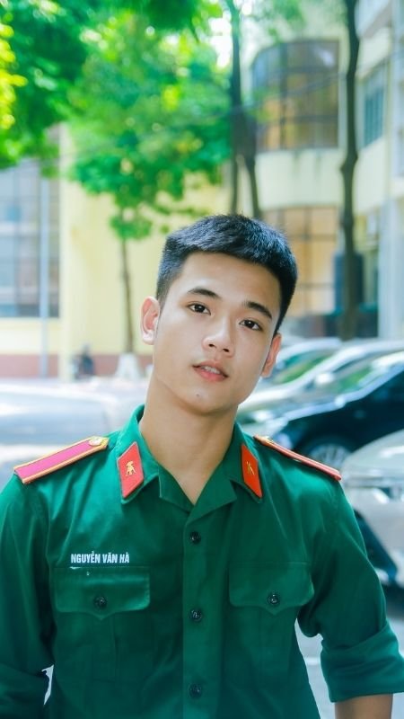 Thanh niên mặc đồng phục quân đội màu xanh đứng trước tòa nhà, xung quanh có cây cối và xe hơi.