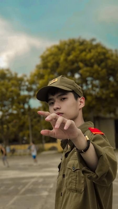 Người mặc quân phục đứng ngoài trời, giơ tay về phía máy ảnh, nền là cây cối và bầu trời.